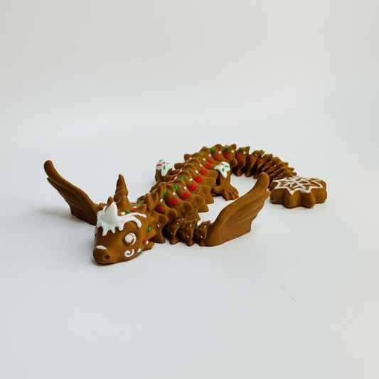 A small brown dragon figurine decorated with white icing-like details and red and green candy-like accents, posed on a plain light background. A snowflake-shaped piece is visible near the tail.