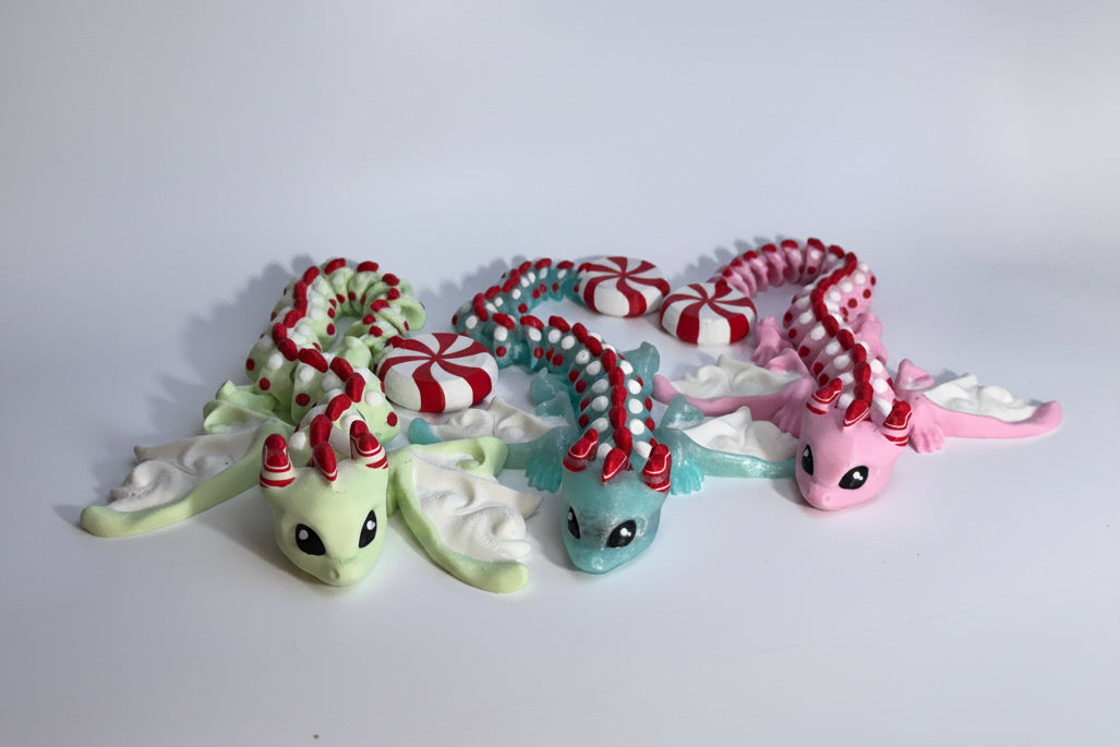Three small dragon figurines arranged side by side on a plain white studio background. The left dragon is pale green with red-and-white striped horns and white wings.