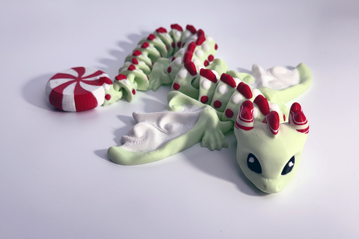 A small dragon figurine in pale green and white with red accents, posed on a plain white background. The dragon has large black eyes, a red-and-white striped horn crown, and a red-and-white striped candy-like wing or accessory near its head.