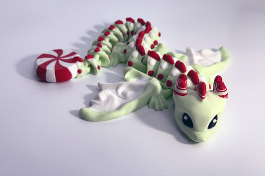 A small dragon figurine in pale green and white with red accents, posed on a plain white background. The dragon has large black eyes, a red-and-white striped horn crown, and a red-and-white striped candy-like wing or accessory near its head.